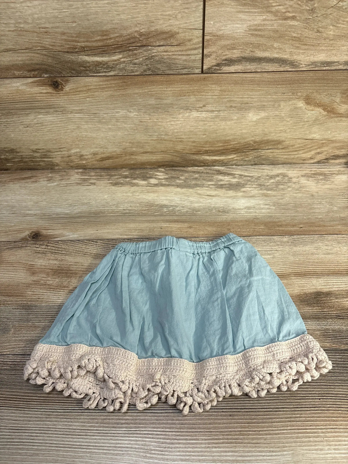 Flat-lay of blue toddler skirt (12-18 months) showcasing elastic waistband and cream crochet pompom hem on wood background