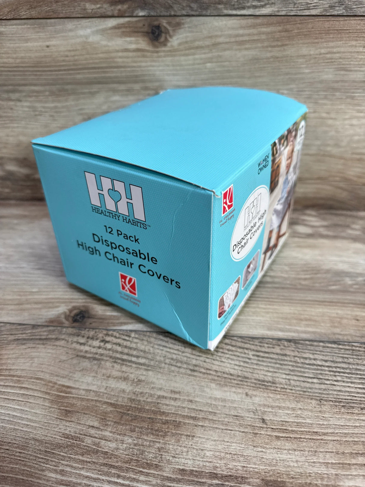 Angled three-quarter view of a 12-pack box of disposable high chair covers showcasing blue packaging and Healthy Habits branding on a wood tabletop