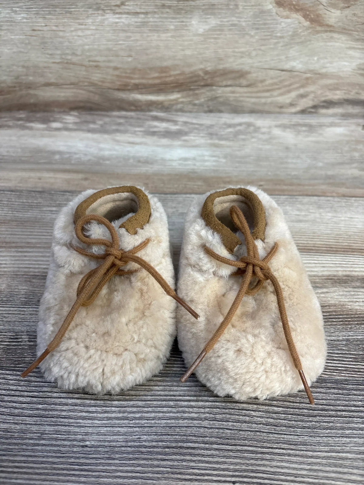 Soft beige shearling baby booties with brown leather laces on rustic wood background