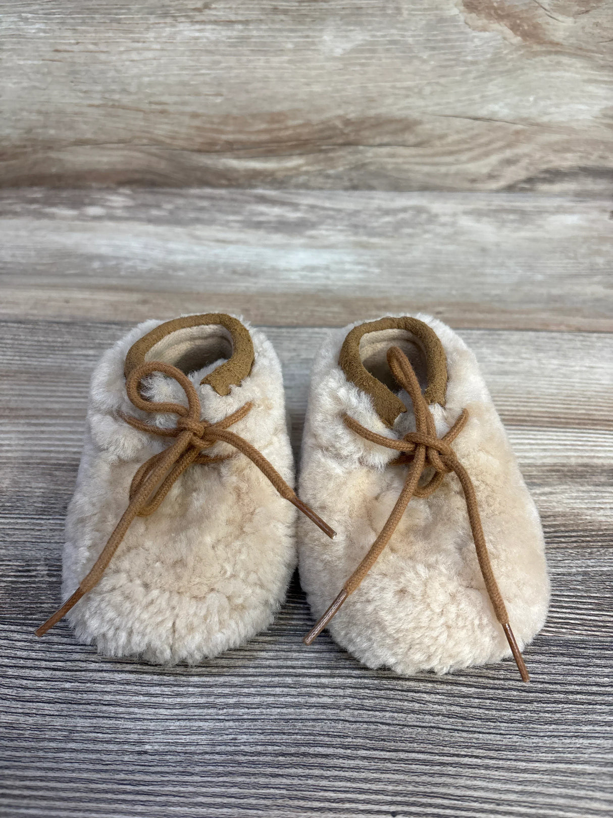 Soft beige shearling baby booties with brown leather laces on rustic wood background