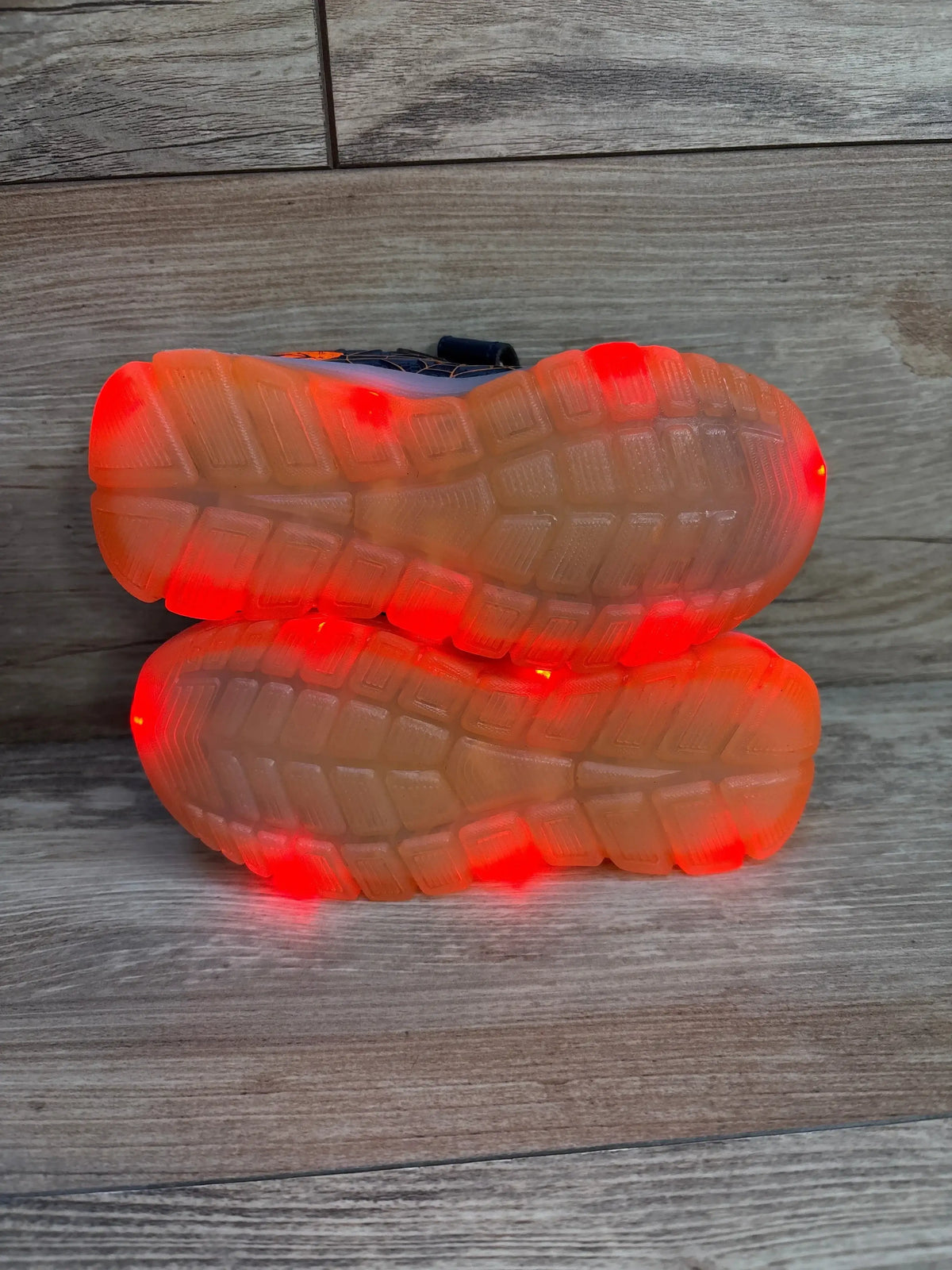 Soles-up view of navy and orange Jian Dian toddler light-up sneakers showing red LED-lit translucent soles on wood floor