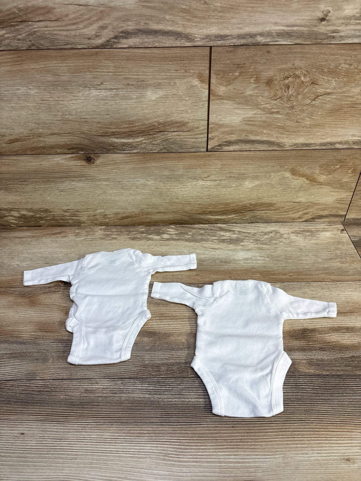Top-down view of two Gerber preemie white long-sleeve bodysuits showing envelope shoulders and snap closures on wood background