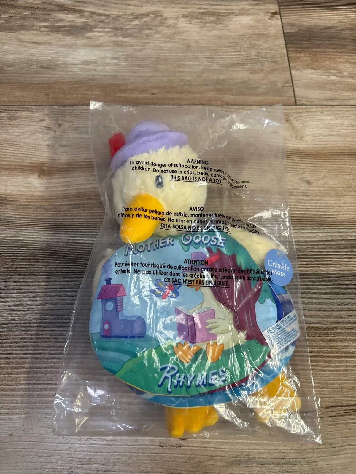 Front view of Mother Goose plush story book with 'Mother Goose Rhymes' cover and crinkle-pages tag, packaged in clear plastic on wood floor