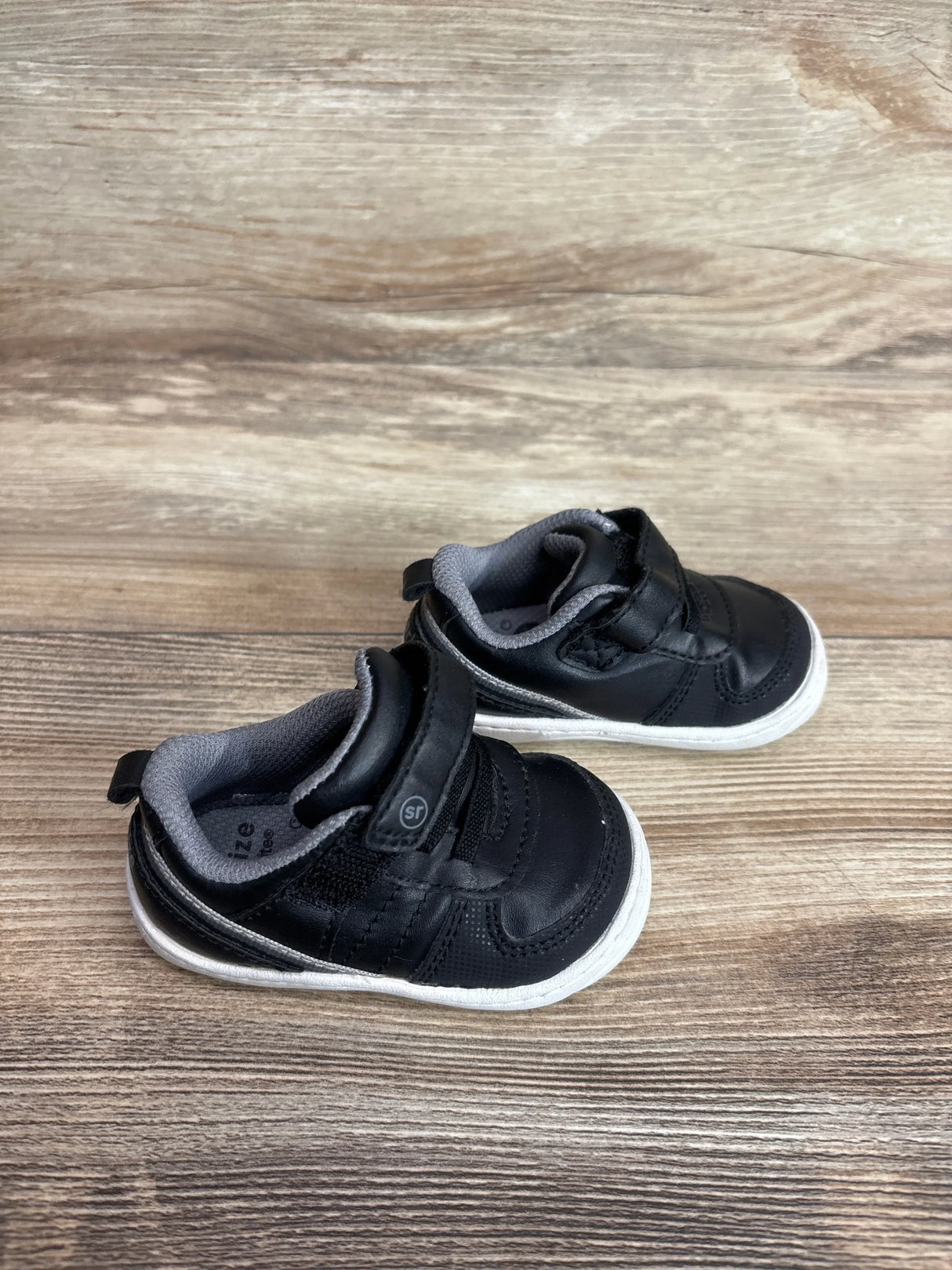 Angled top view of Surprize by Stride Rite black toddler sneakers showing Velcro straps and white soles on wood background