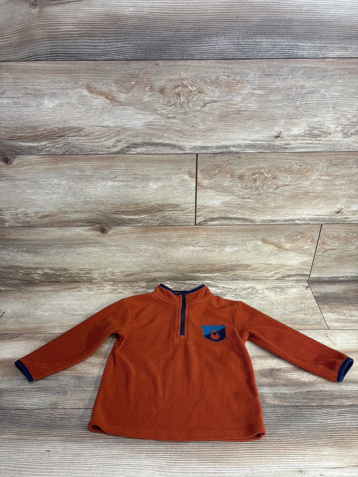 Orange fleece long sleeve kids pullover with blue bear patch and half zip, laid flat on wood