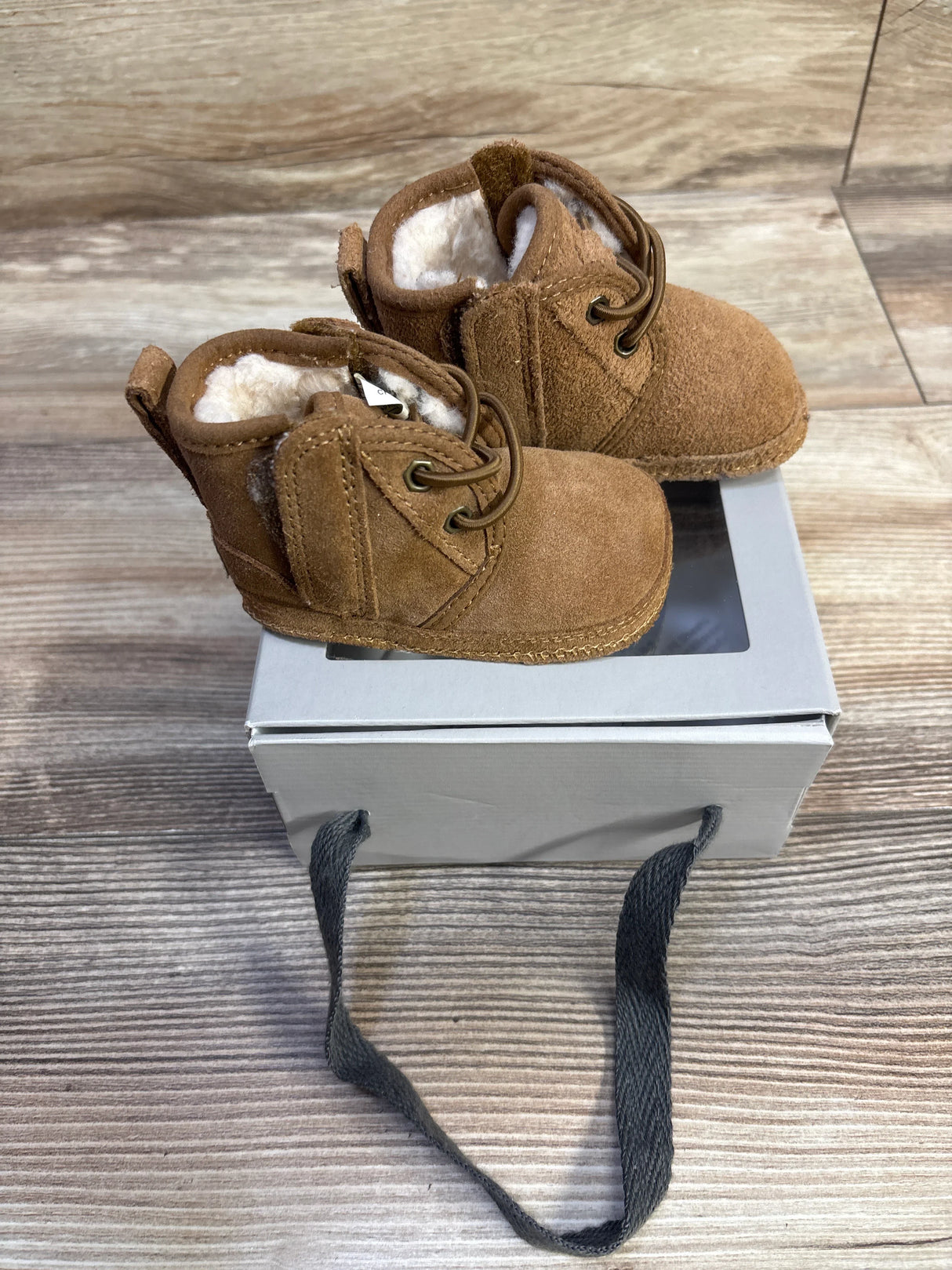 Top-front view of Ugg Baby Neumel chestnut booties showcasing suede upper, faux-fur lining, and laces on a gray box and wood floor