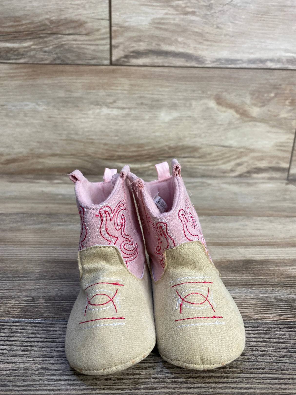 Front view of Stepping Stones baby cowgirl crib boots in pink and tan, embroidered upper and suede-look toe on wood floor