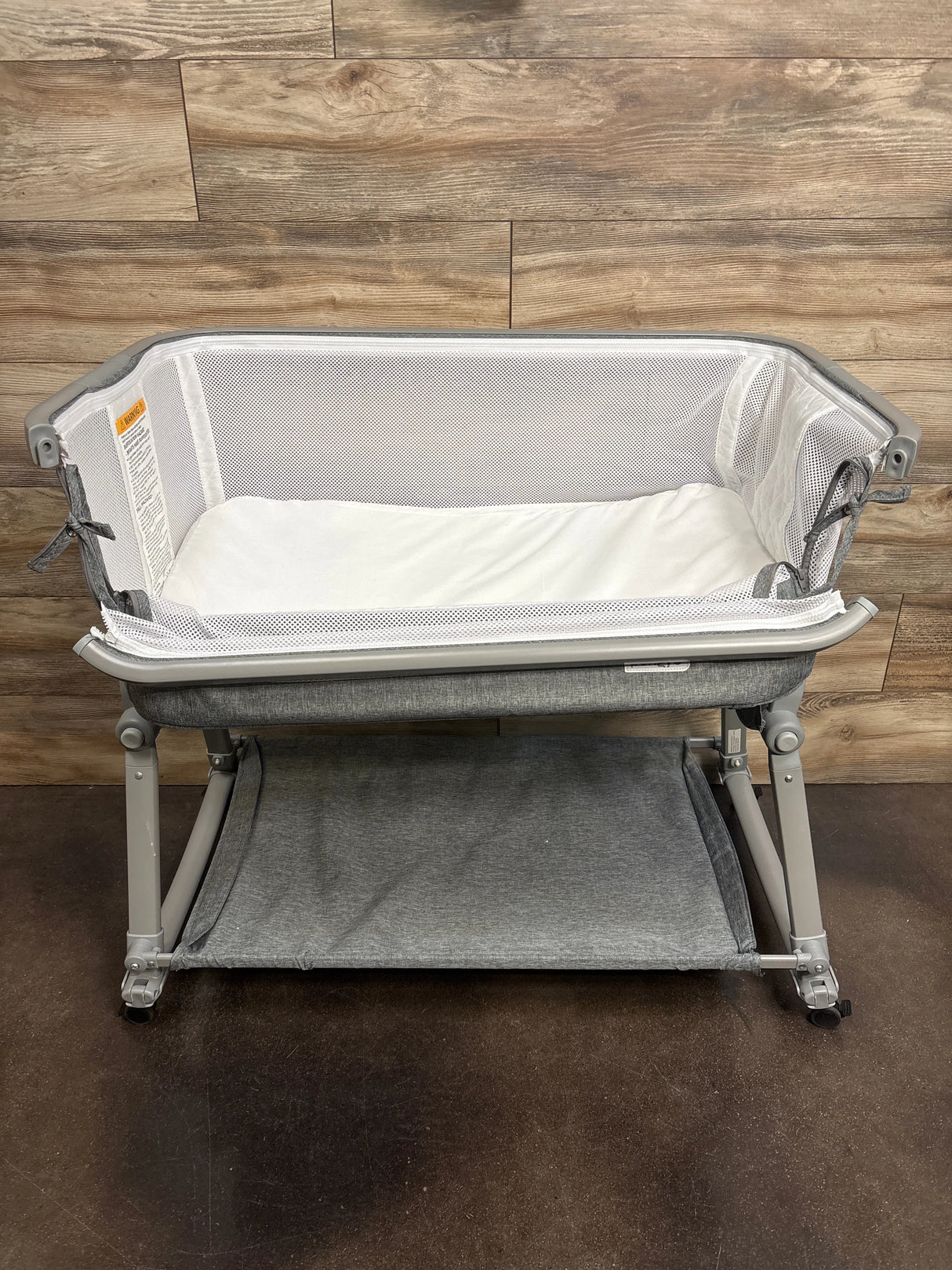 Gray portable baby bassinet with mesh sides and storage shelf on wood floor