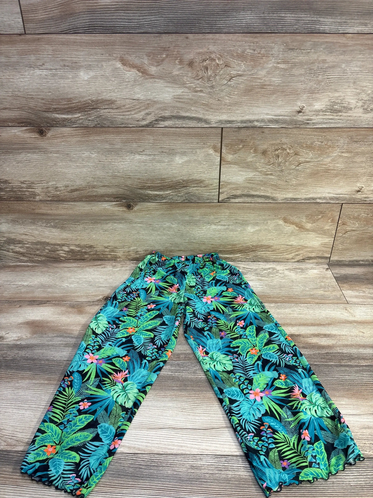 Flat lay of teal tropical print swim cover up pants showing elastic waistband and ruffled ankle hems on wood background
