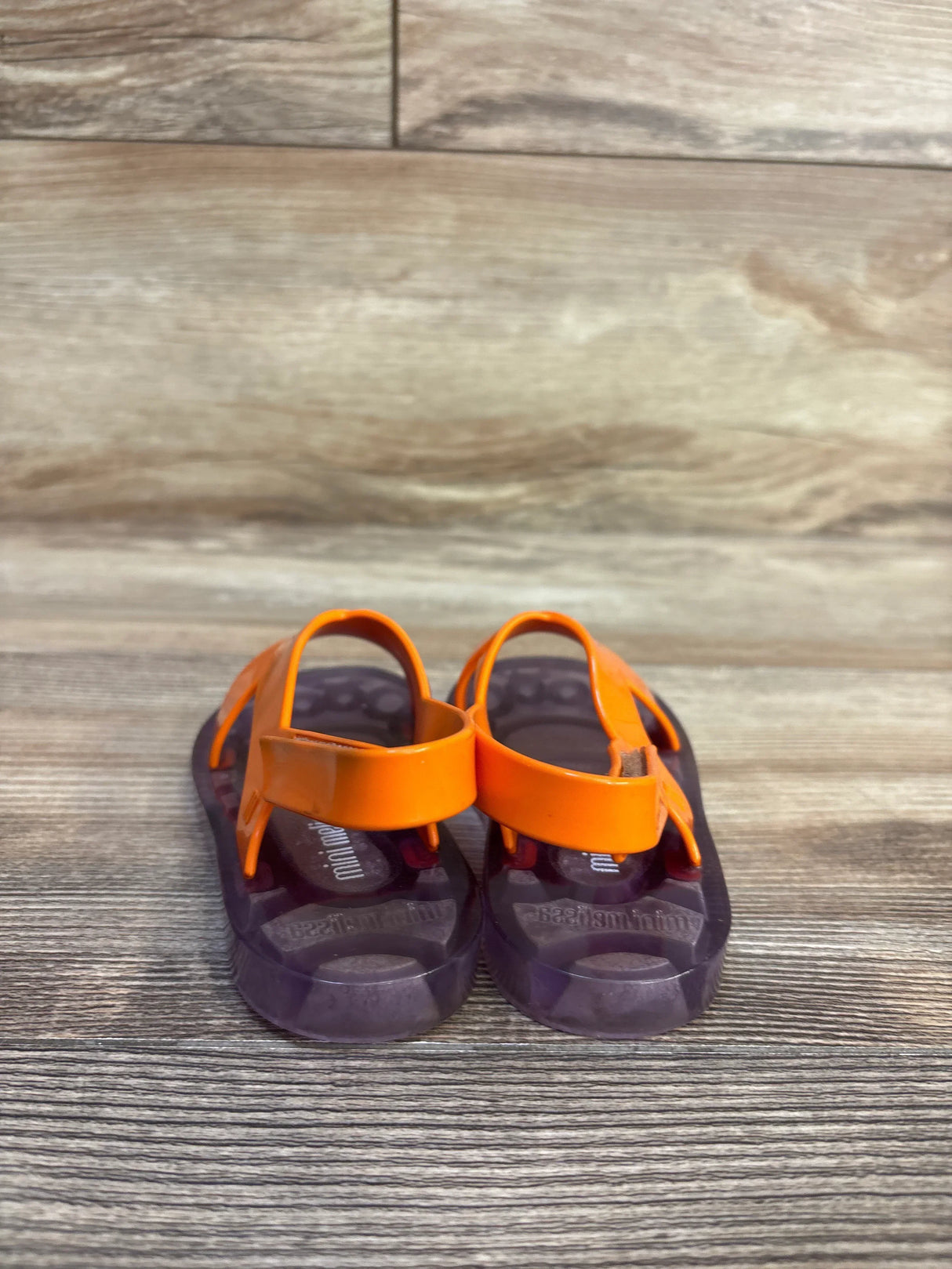 Back view of Mini Melissa Friendly BB jelly sandals, 8C, with orange straps and purple soles on wood background