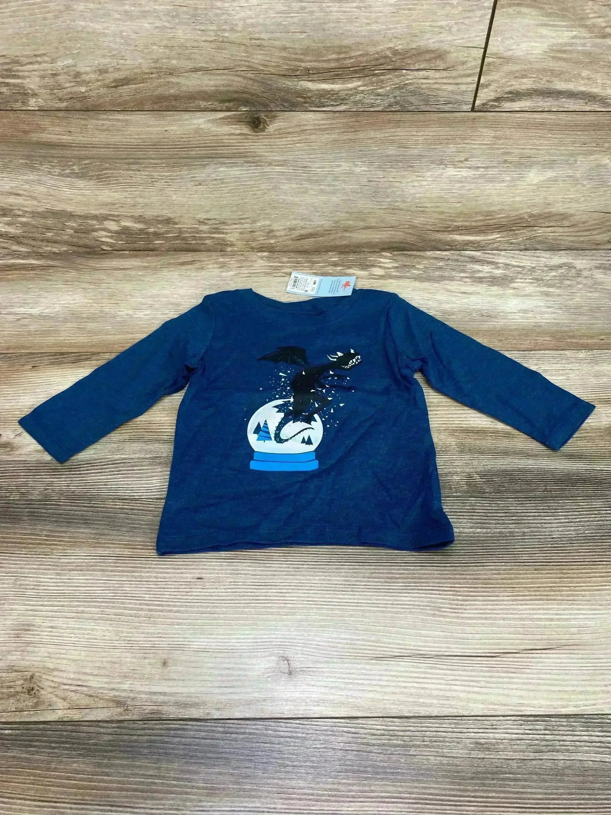 Front flat-lay of Cat & Jack 12M long-sleeve baby shirt with black dragon snow-globe print on blue, wood background