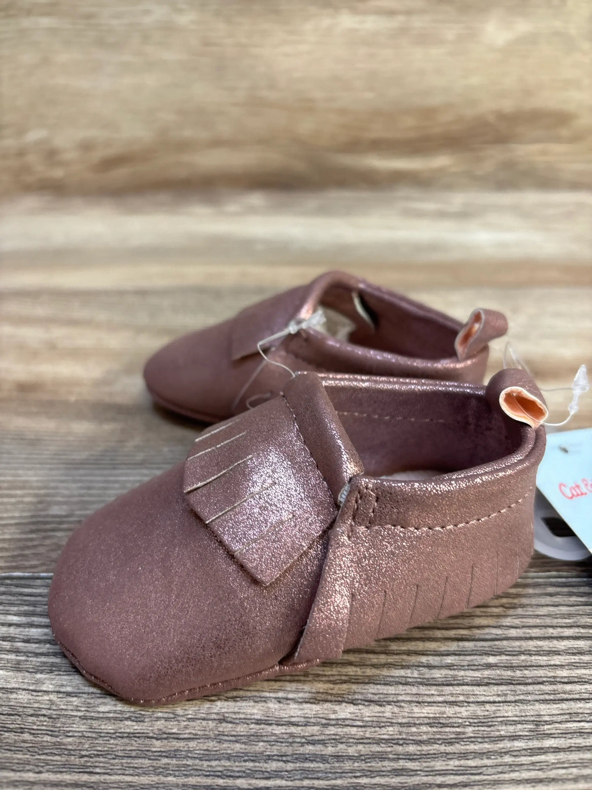 Metallic pink baby moccasins with fringe detail on wooden surface