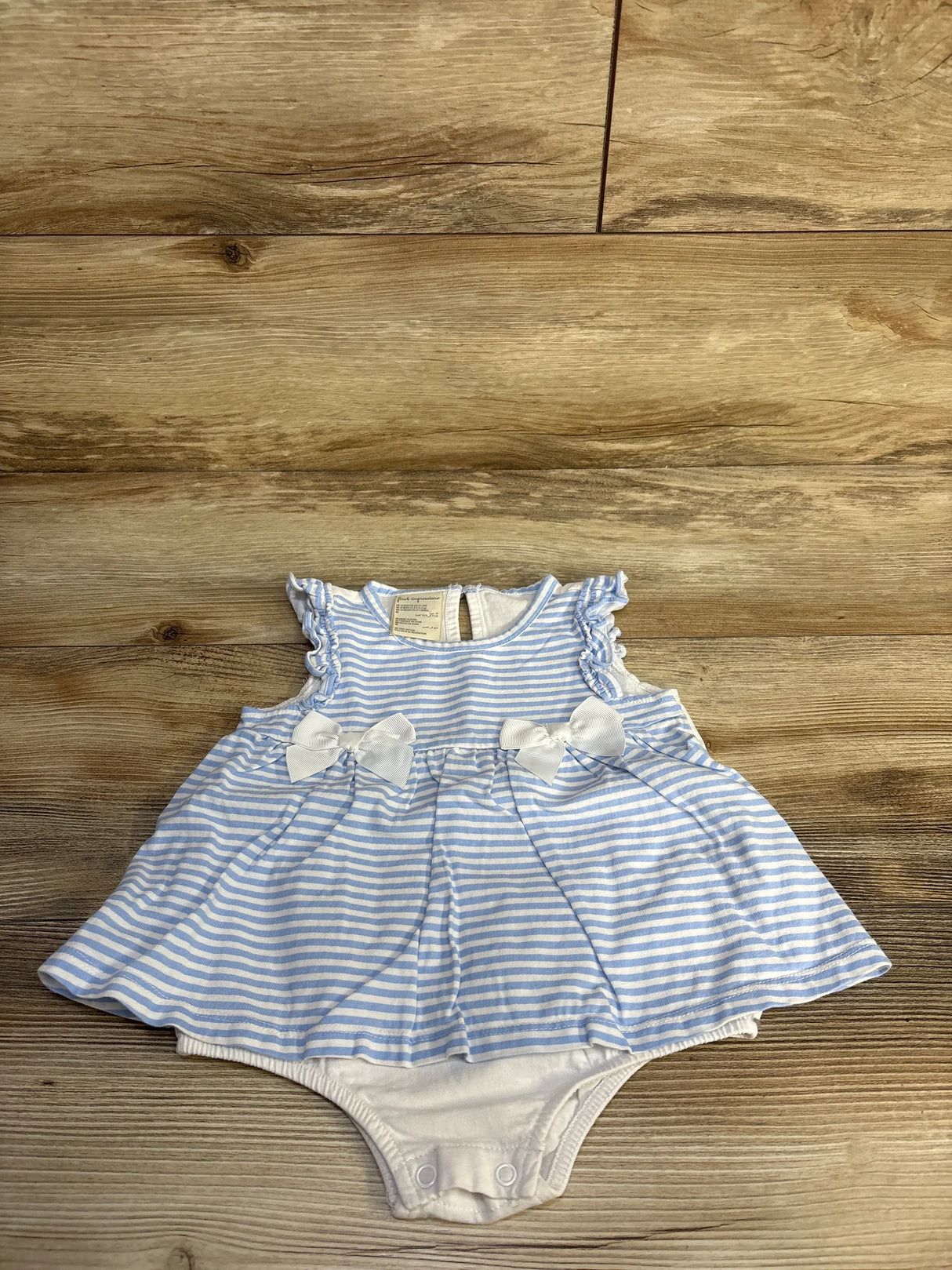 Flat-lay of blue and white striped baby bodysuit dress showcasing ruffle cap sleeves, two front bows, gathered skirt and snap-closure crotch on wood background