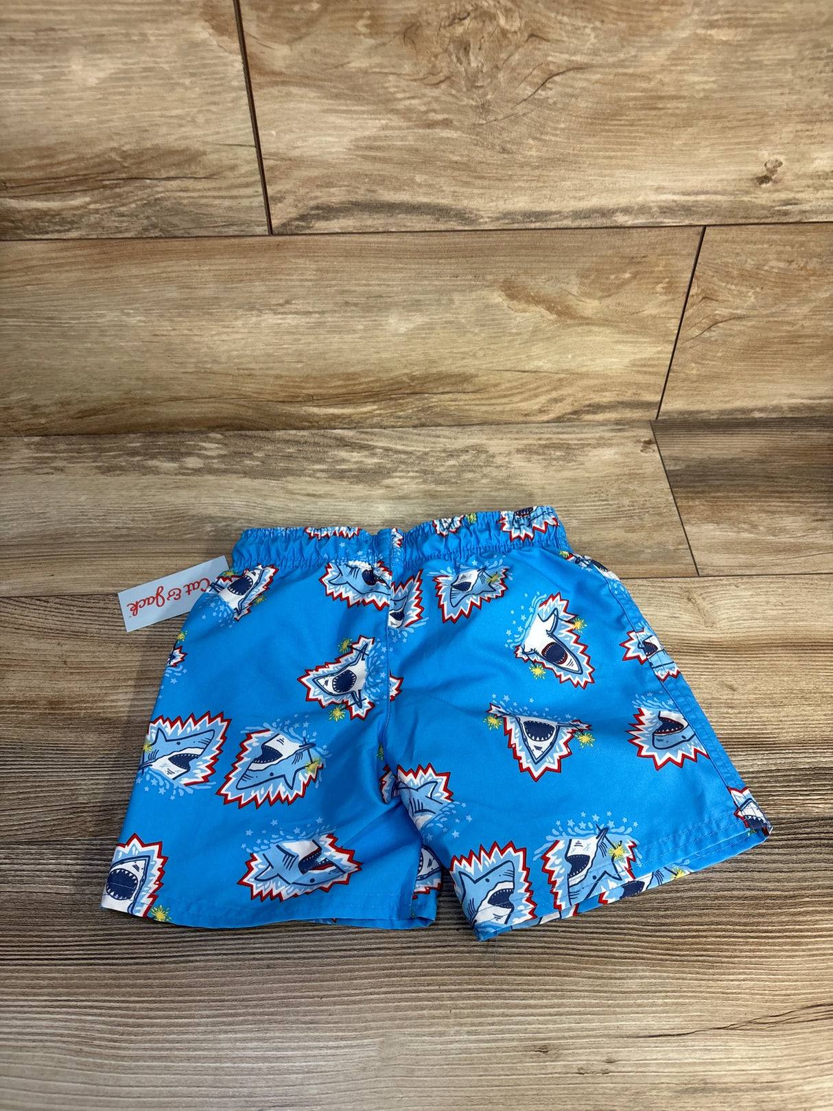 Flat-lay of Cat & Jack blue toddler swim trunks showing elastic waistband and sharks with sparklers print on wood background