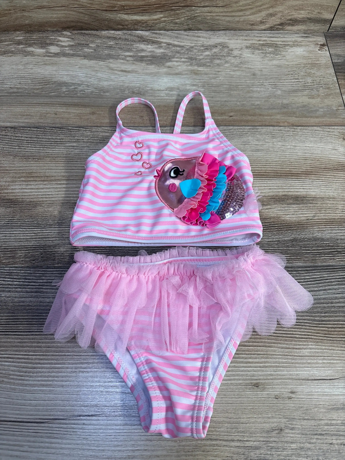 Flat lay of pink and white striped 2-piece toddler swimsuit showing bird applique with sequins and tutu skirt on wood background