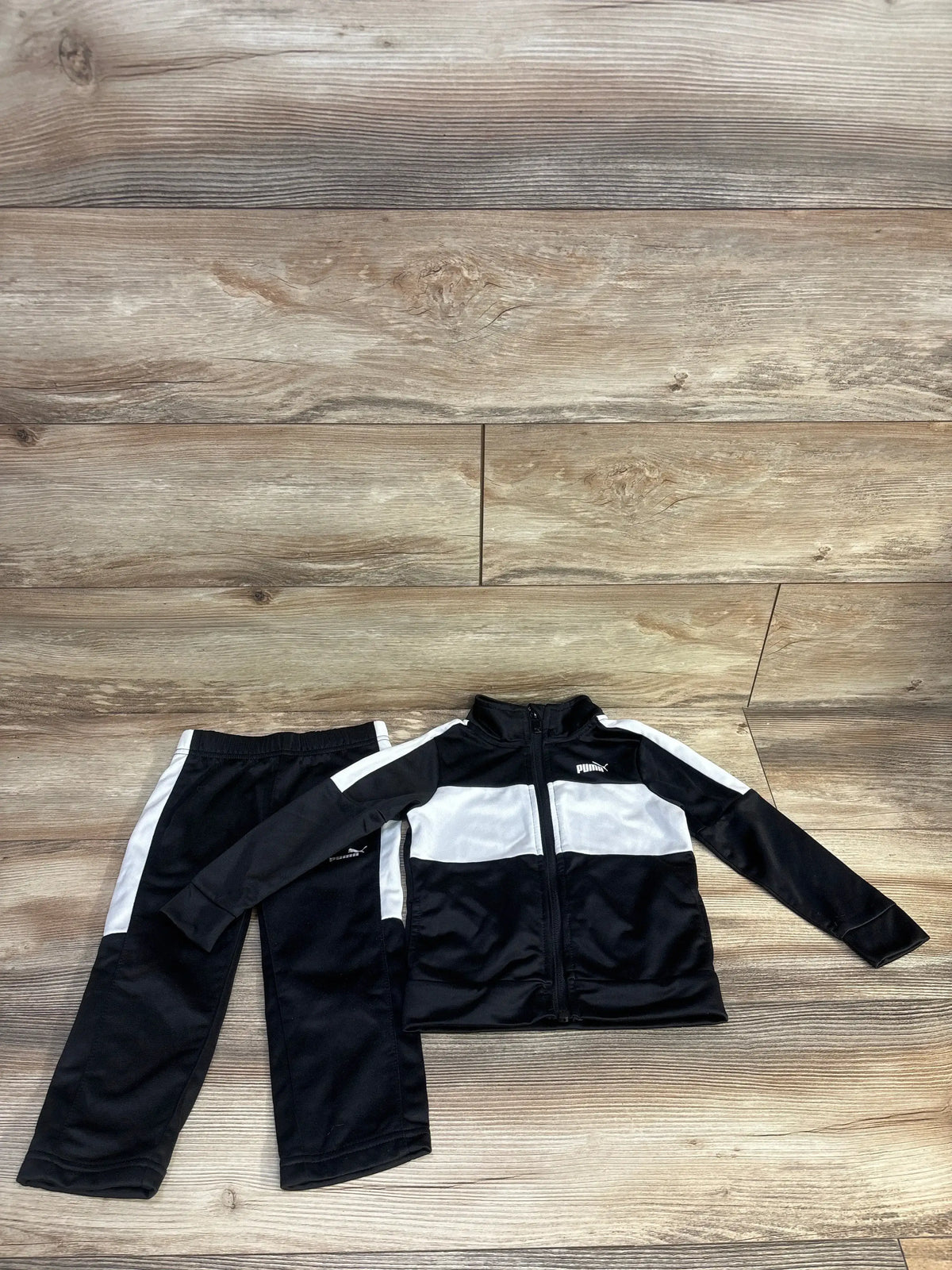 Flat lay of toddler Puma 2-piece tracksuit showing zip-up jacket with white chest panel and matching pants on wood background