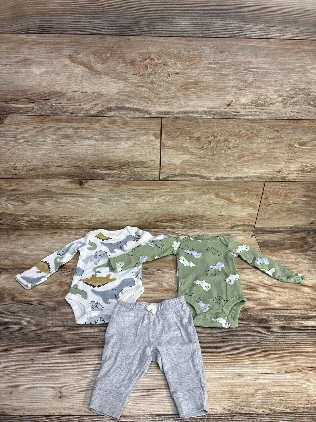 Overhead view of Carter’s three piece bodysuit set in green and gray with dinosaur print bodysuits and gray pants on wood flooring