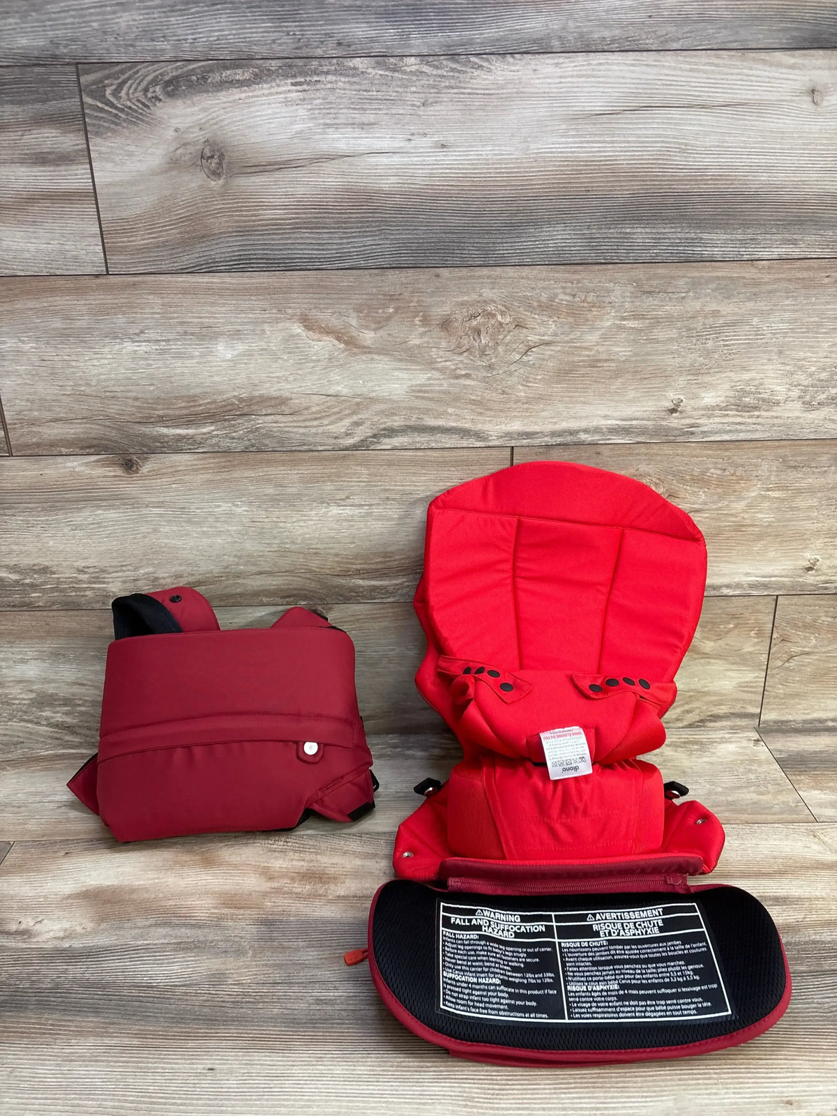 Front of Diono Carus baby carrier in red with padded shoulder straps, detachable waist and visible safety label on wood floor