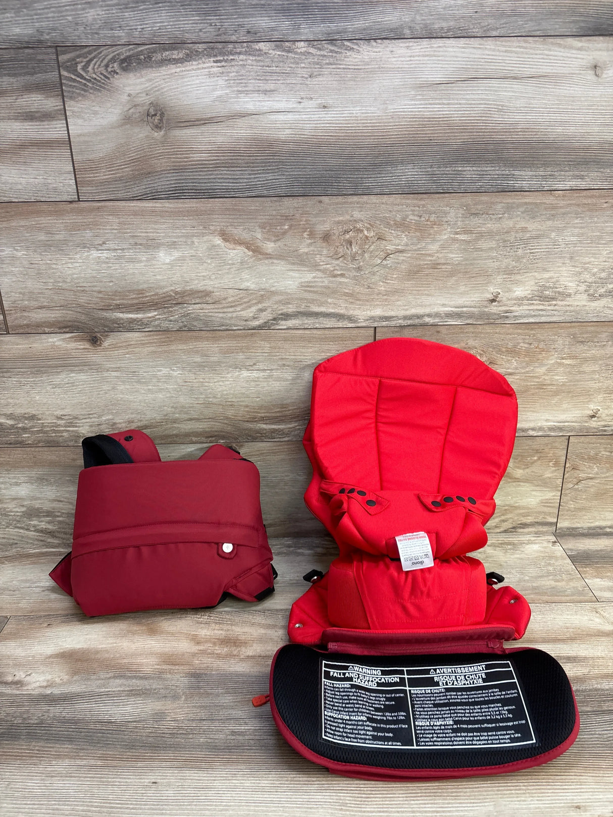 Red baby carrier with padded straps and safety label displayed on wood floor