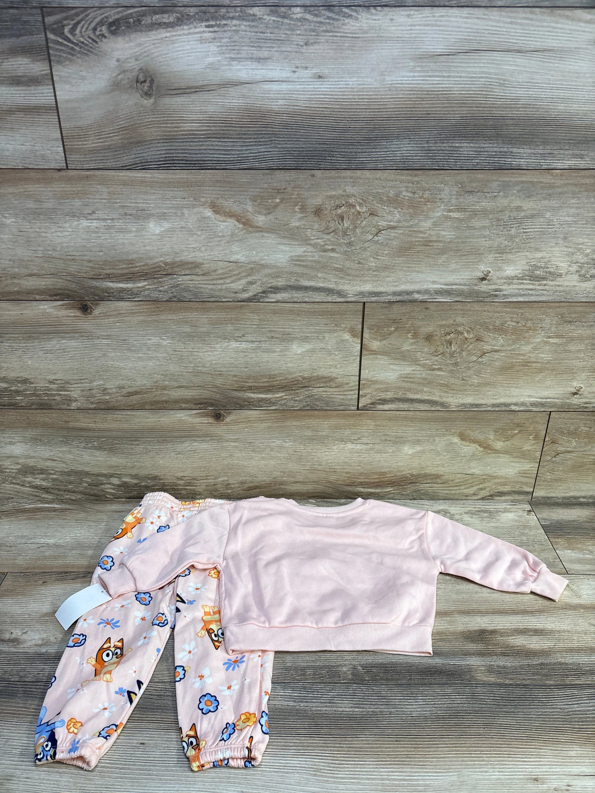 Flat lay of pink Bluey 2-piece sweatshirt and joggers showcasing a bow on the sweatshirt and Bluey floral-print joggers on a wood floor