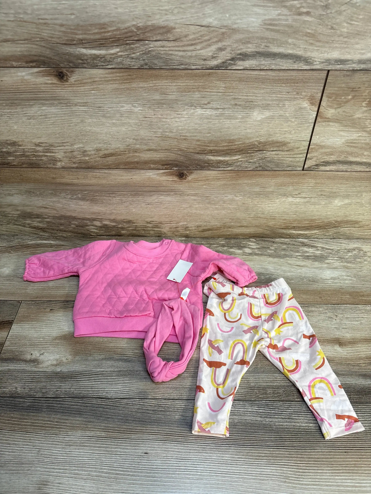 Flat lay of pink quilted baby sweatshirt with matching headband and pink rainbow print leggings on wood plank floor