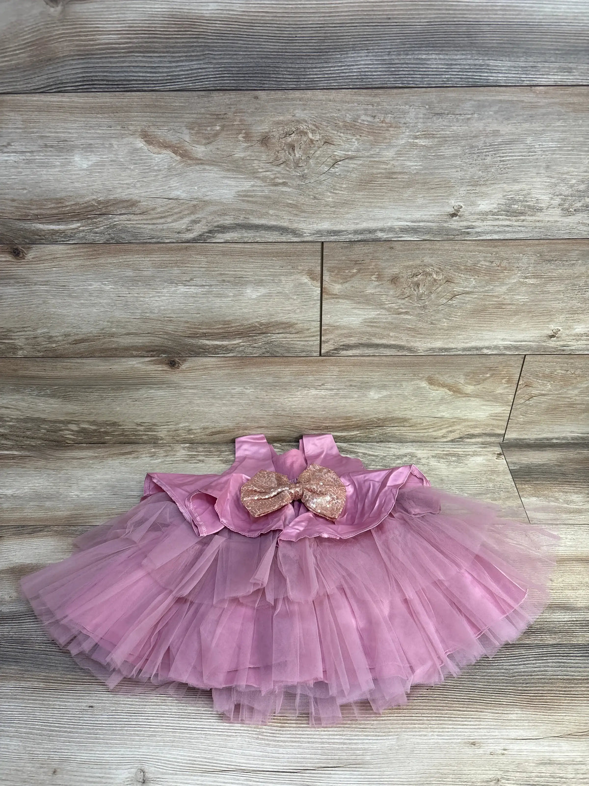 Flat lay of pink bowknot satin dress showing layered tulle skirt and sequin bow on light wood background, size 9-12 months