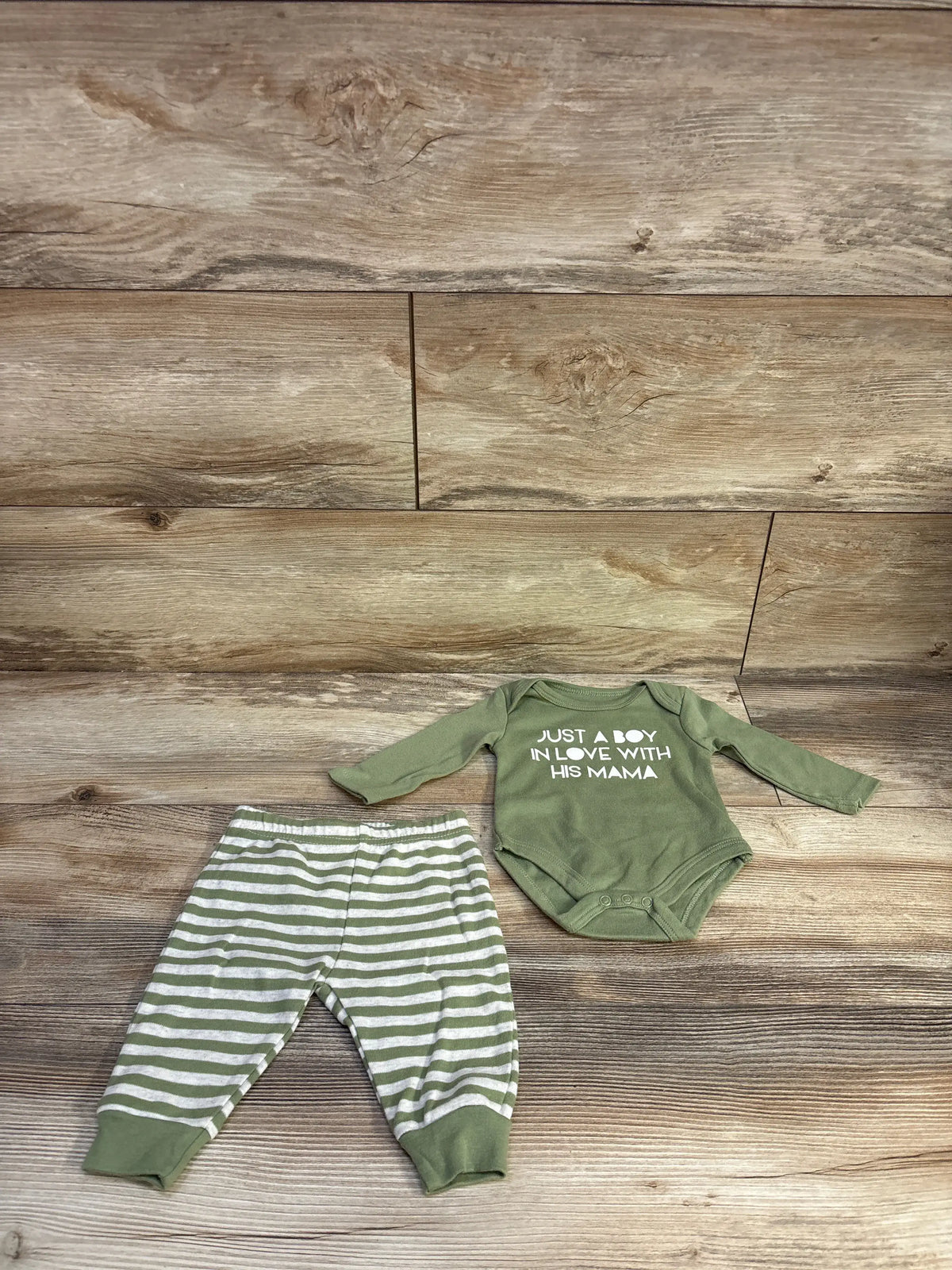 Flat lay of green 2-piece baby set with long-sleeve bodysuit reading Just a boy in love with his mama and green striped pants on wood plank backdrop