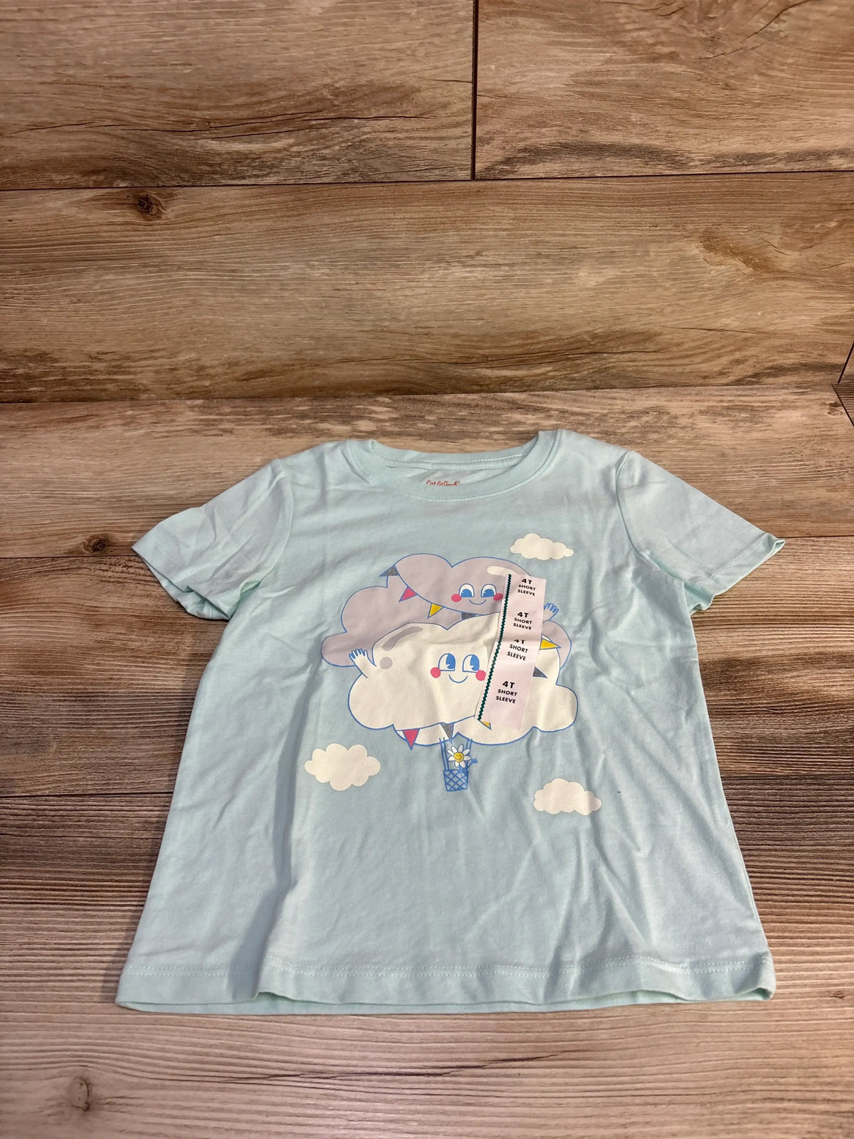 Front flat lay of light blue Cat and Jack 4T short sleeve toddler shirt showing smiling cloud graphic and small clouds on wood background