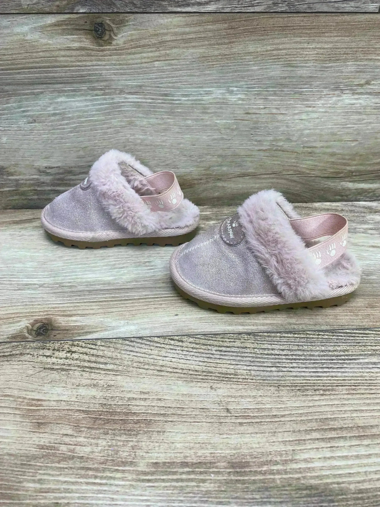 Side view of BEARPAW Rebecca suede toddler slippers showing fluffy sheepskin lining and elastic heel strap on wood background