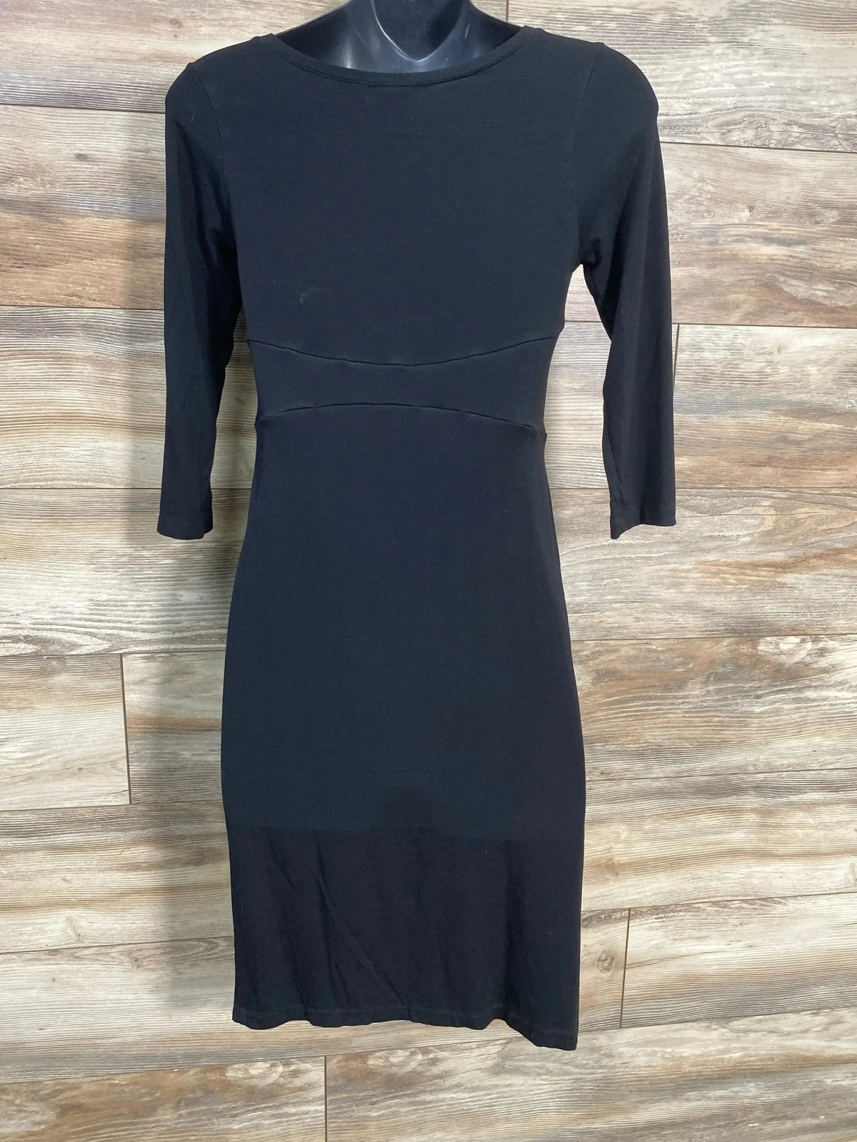 Front view of Isabella Oliver dress, black 3/4 sleeves, fitted empire waist and knee-length skirt on mannequin, wood backdrop