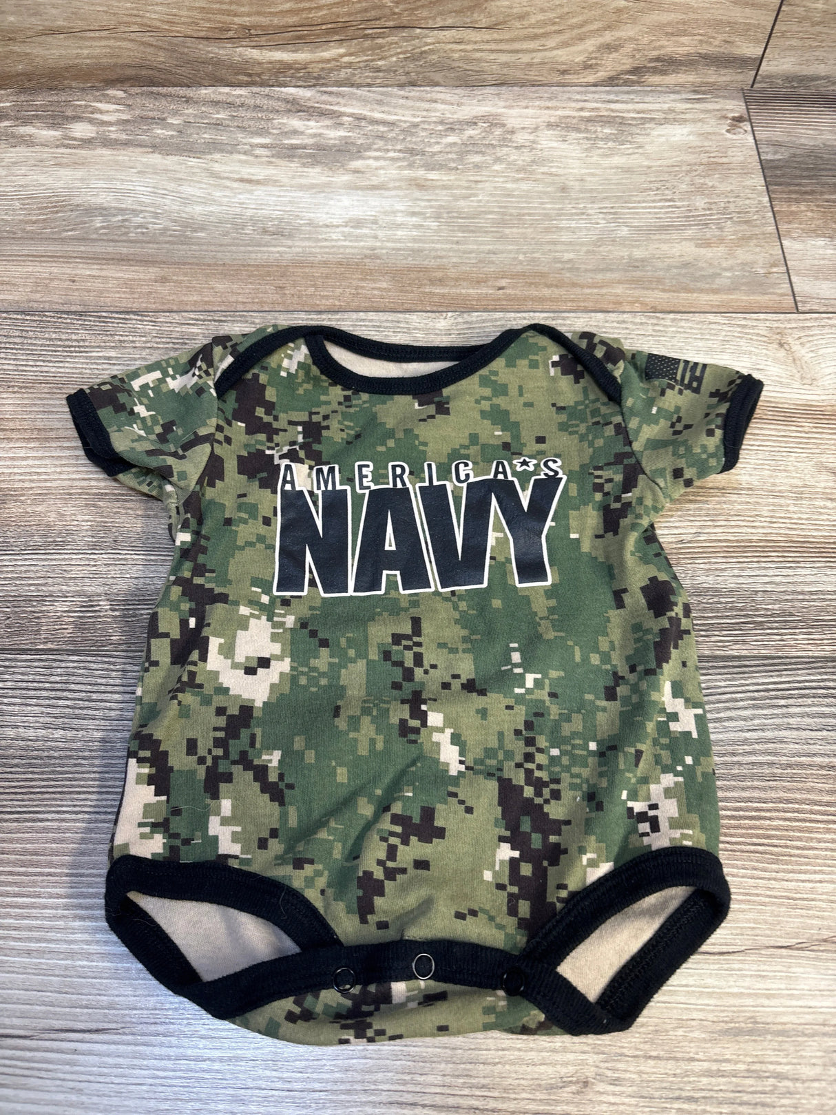 Green camo baby onesie with 'America's Navy' text, short sleeves, snap closure, on wood floor.