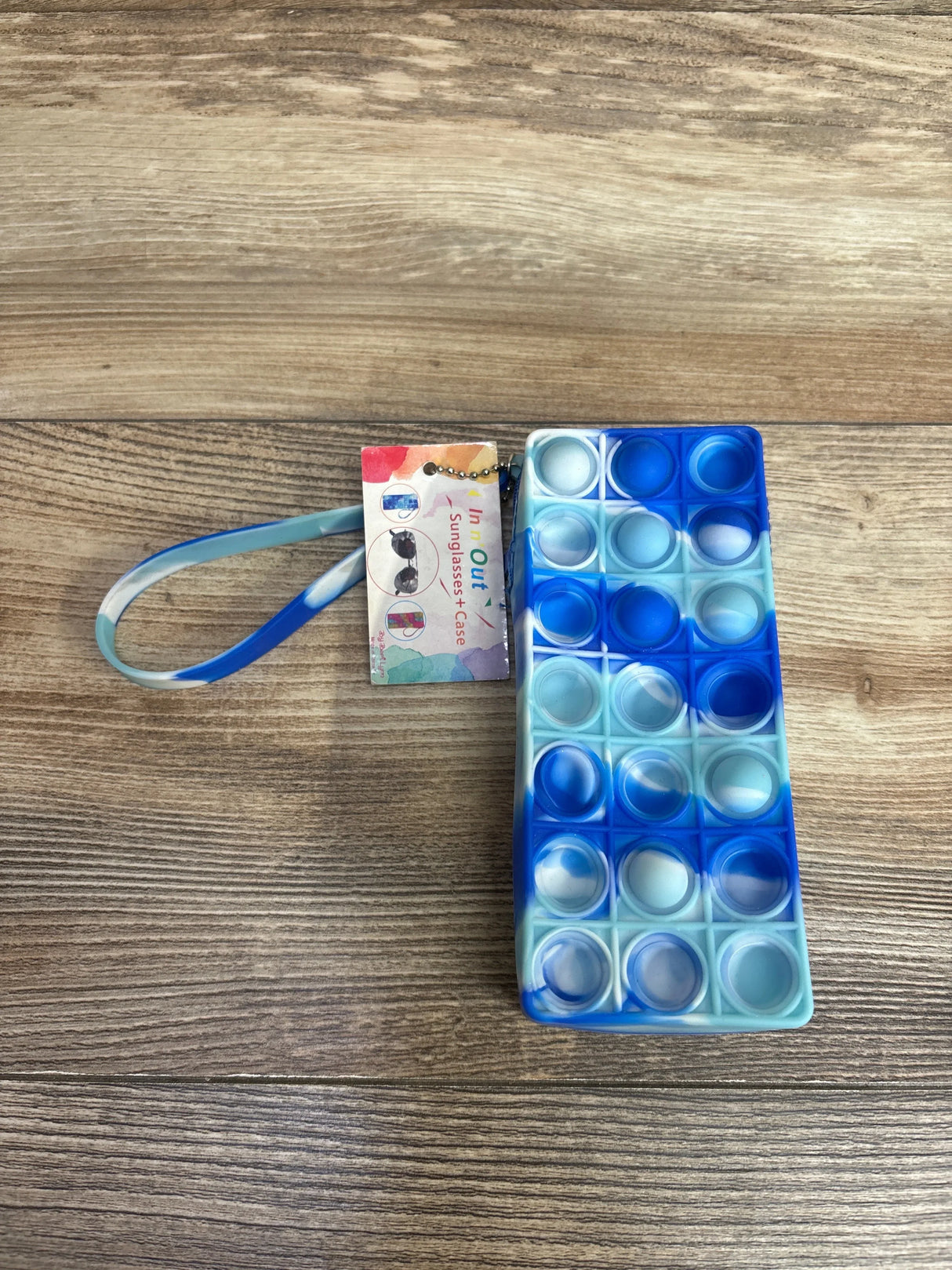 Top-down of Pop It In N Out Sunglass Case Blue Marble showing rectangular silicone pop-it bubbles, wrist strap and tag on wood