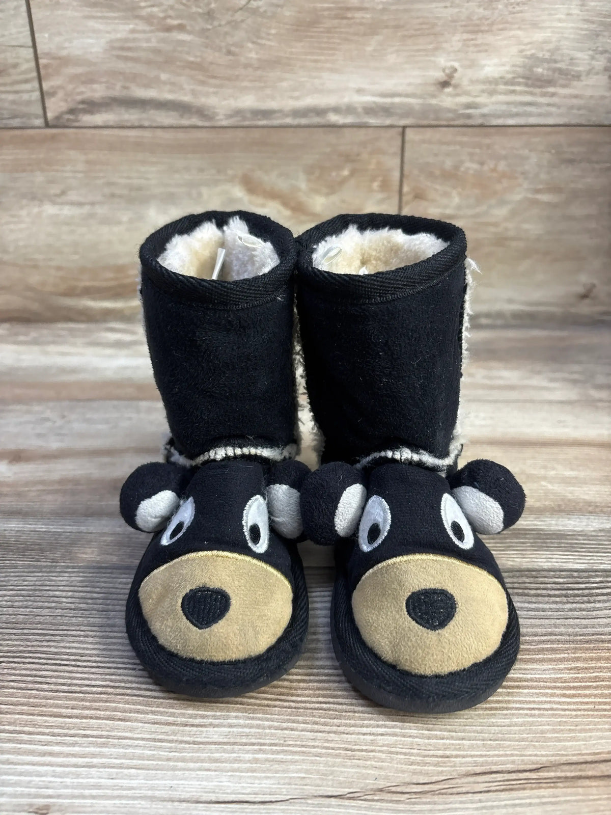 Front view of black kid’s bear slipper boots size 6-7C showing plush bear faces and cream faux-fur lining on wood background