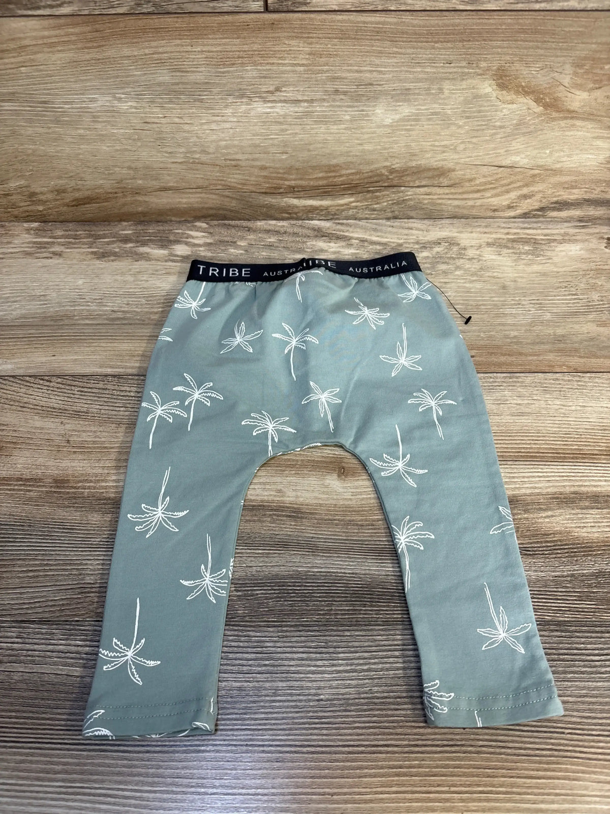 Green baby pants with white palm tree print and black waistband, laid on wood floor