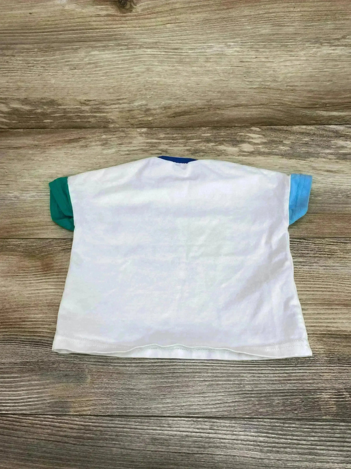 Back view of Mayoral dino pocket shirt sz 2-4m showcasing white body with teal and blue color-block sleeves on wood plank floor