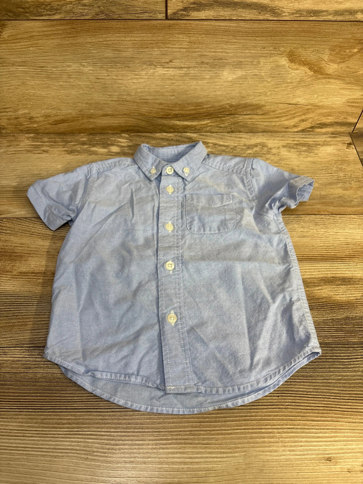 Flat lay of blue Children's Place short-sleeve button-down shirt showing front buttons and chest pocket on wood background