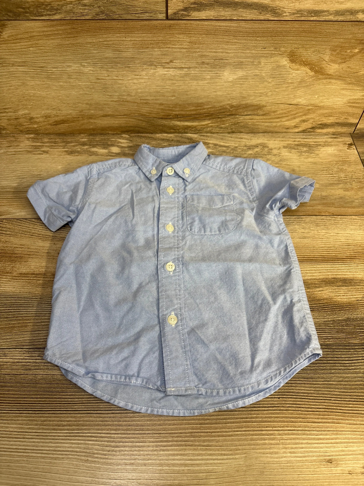 Flat lay of blue Children's Place short-sleeve button-down shirt showing front buttons and chest pocket on wood background