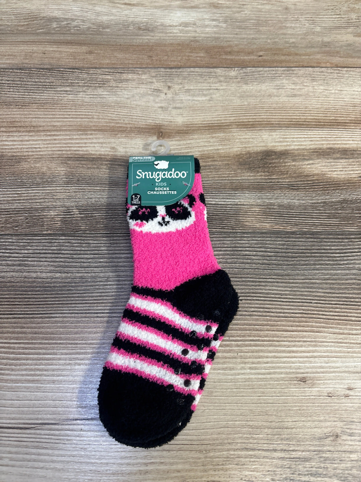 Top-down view of a Snugadoo kids fuzzy pink sock showcasing black toe and heel, white and black stripes, and silicone grip dots with a wood background