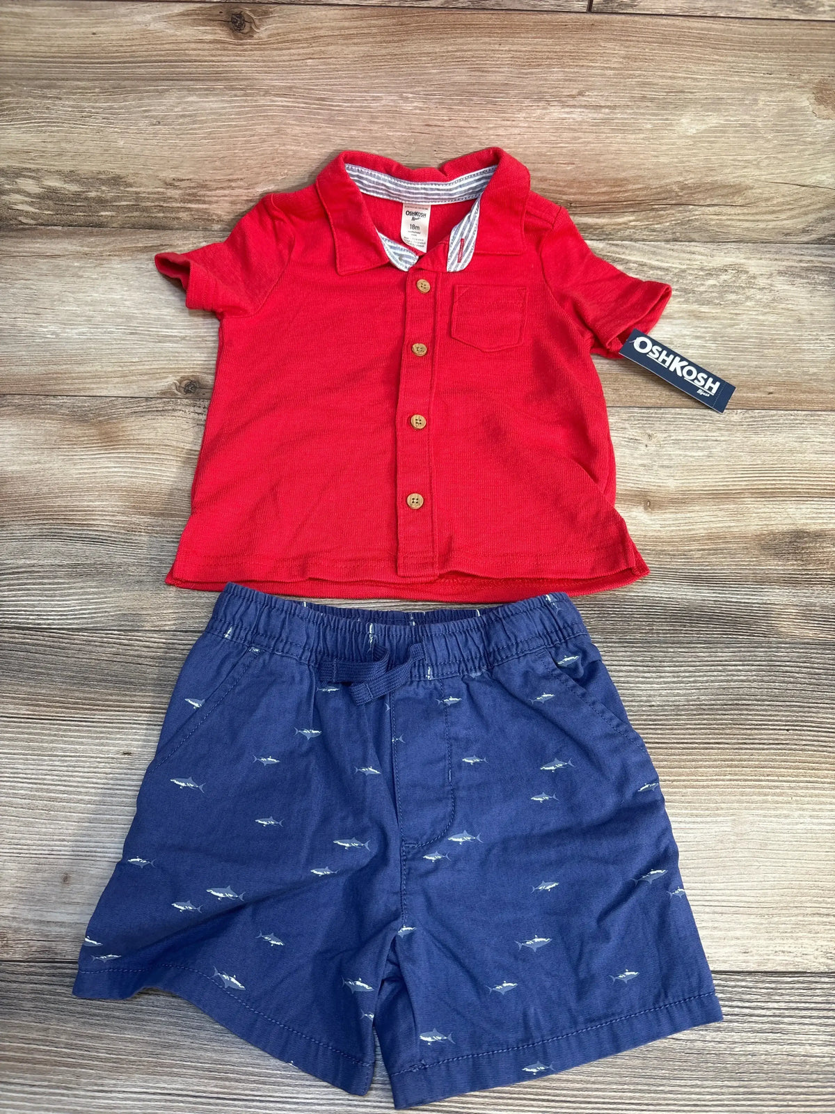 Flat lay of OshKosh button down top and shorts set showcasing red collared shirt and navy shark print shorts with brand tag on wood floor