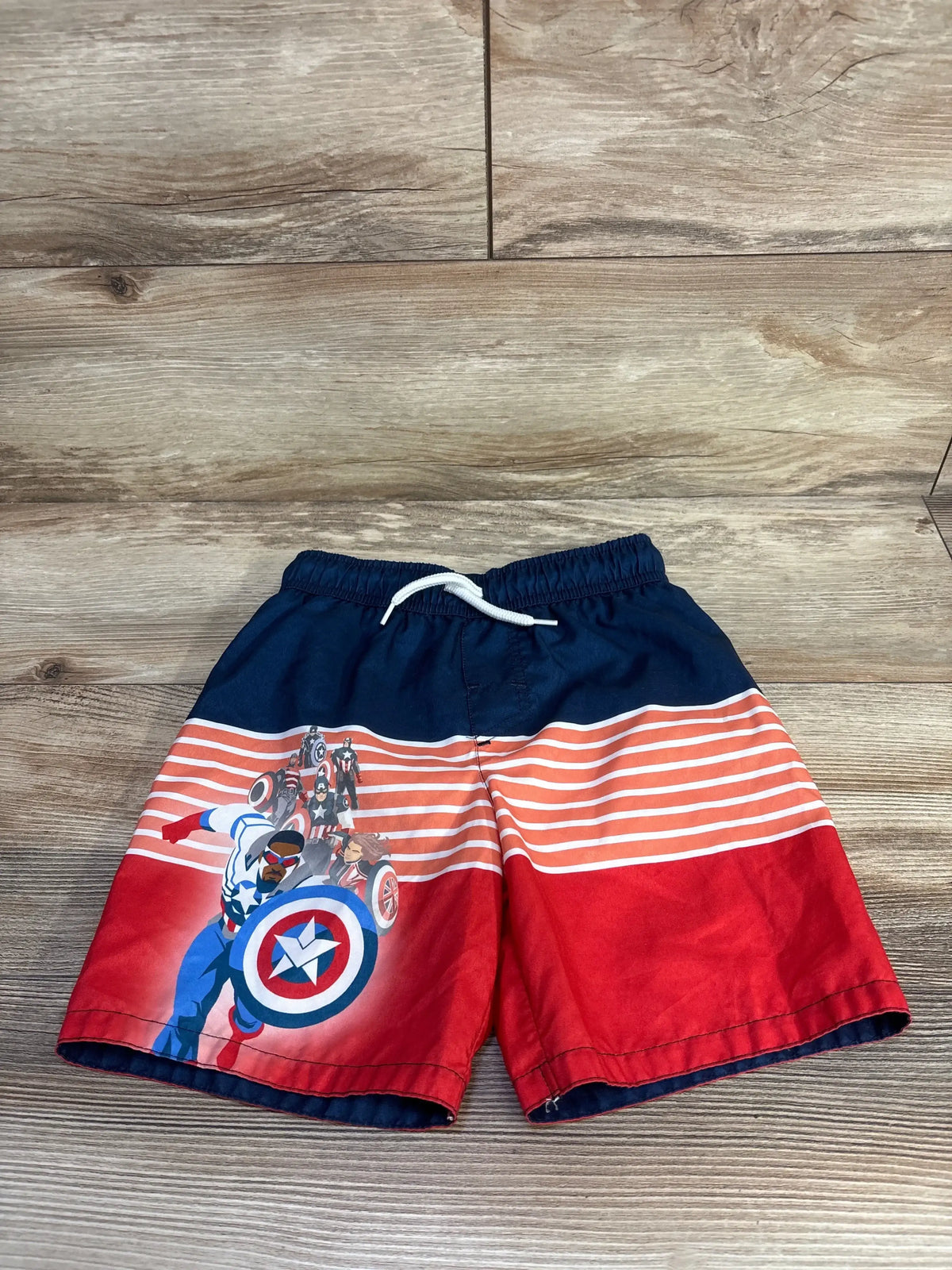 Front view of blue Captain America swim trunks, size 5/6T, with red and white stripes and drawstring on wood background
