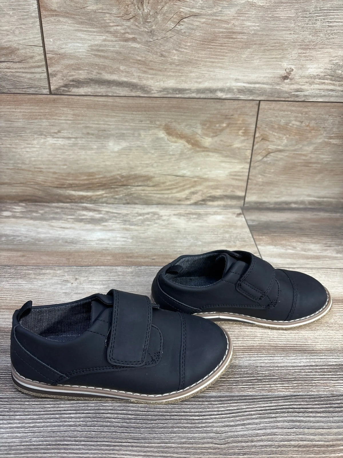 Angled side view of Cat and Jack Adonis black derby shoes showing Velcro strap and stitched contrast sole on wood floor