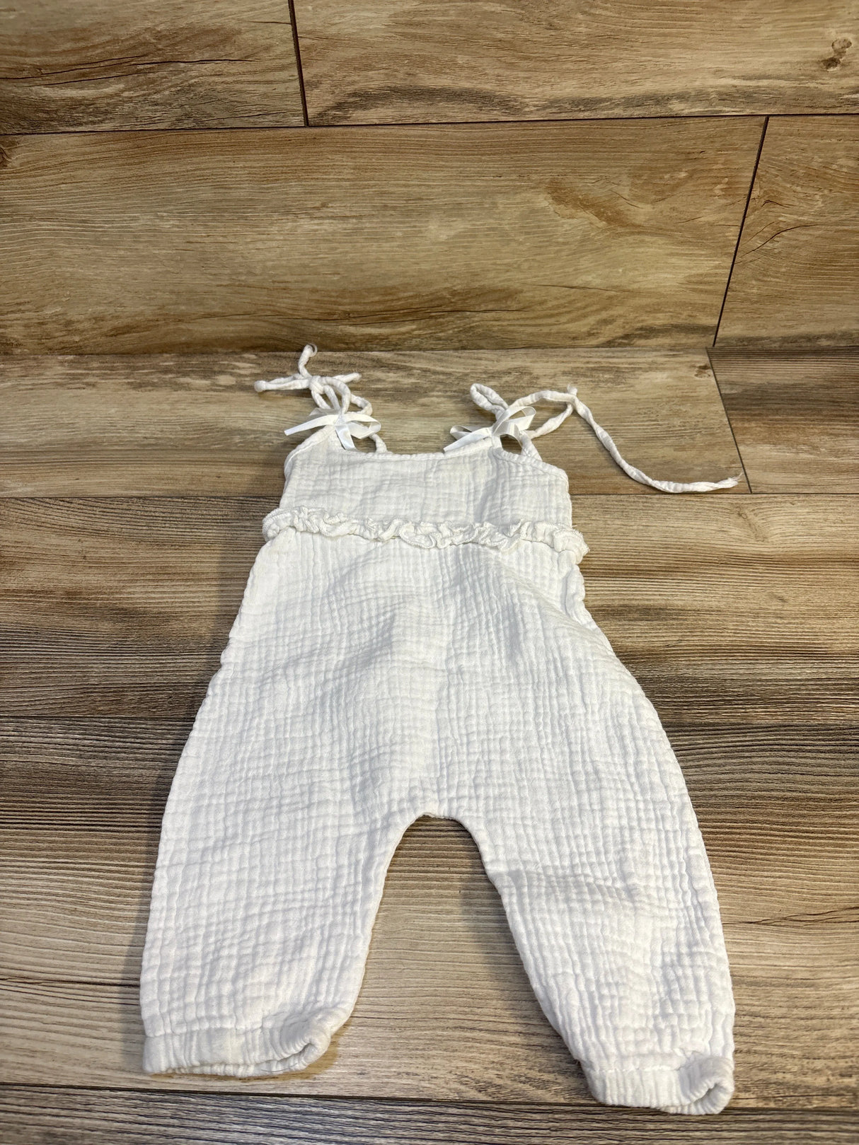 Flat lay of white muslin tank jumpsuit showing tie shoulder straps, ruffle waist, and crinkled gauze on wood background