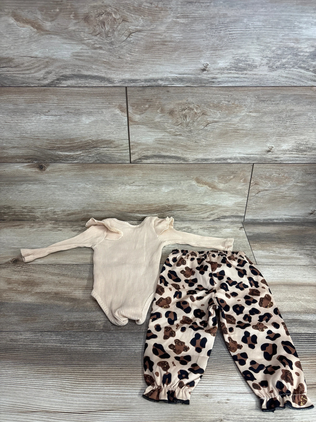 Top-down view of tan ribbed baby bodysuit with ruffle shoulders and matching leopard-print pants on wood floor, size 3-6m