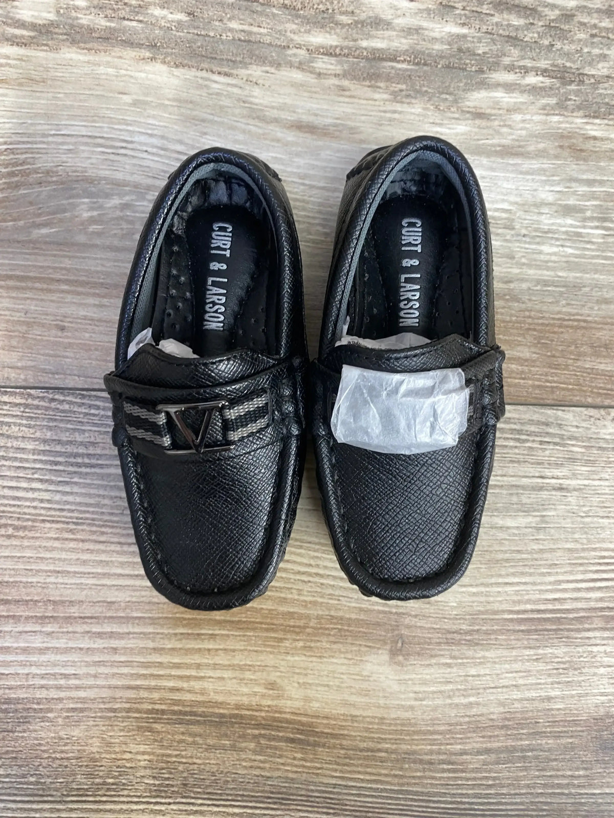 Top view of Curt & Larson black toddler loafers size 5C with textured faux leather and metal V emblem on wood floor