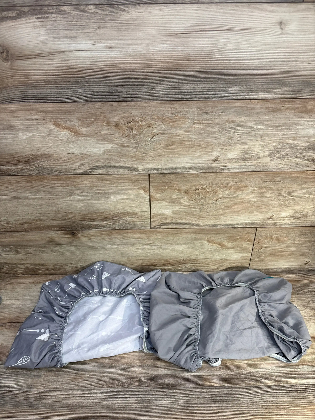 Top-down view of two Biloban fitted bassinet sheets, one solid grey and one grey leaf print, showing elastic edges on wood background