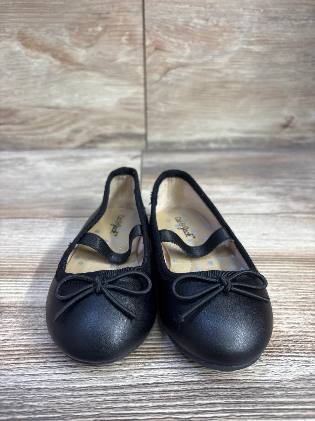 Front view of Cat and Jack Nora slip-on black ballet flats showing bow detail and elastic instep strap on light wood floor