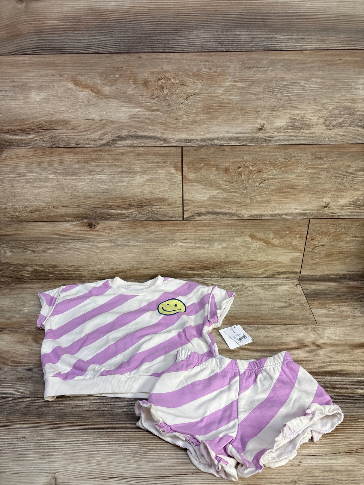 Flat lay of 2-piece striped shirt and ruffle shorts in lavender and white with yellow smiley patch, size 4T on wood