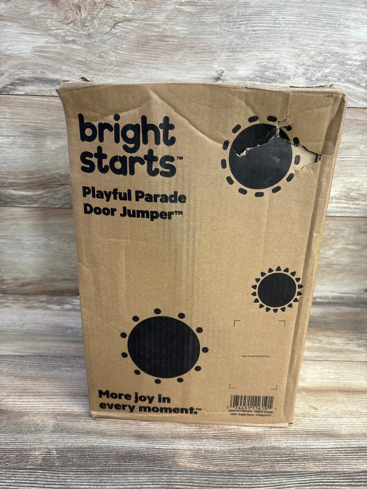 Front view of Bright Starts Playful Parade Door Jumper box with brand logo and tear at top right on wood background