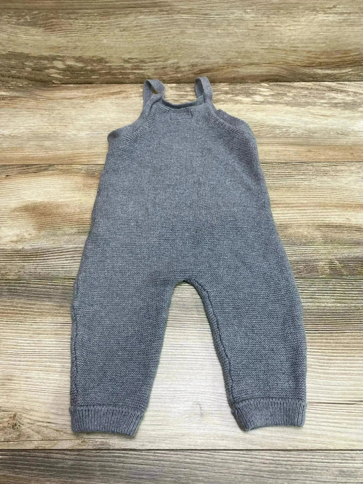Flat lay of Little Planet knit overalls in grey, size 12m, showing shoulder straps and knit texture on wood background