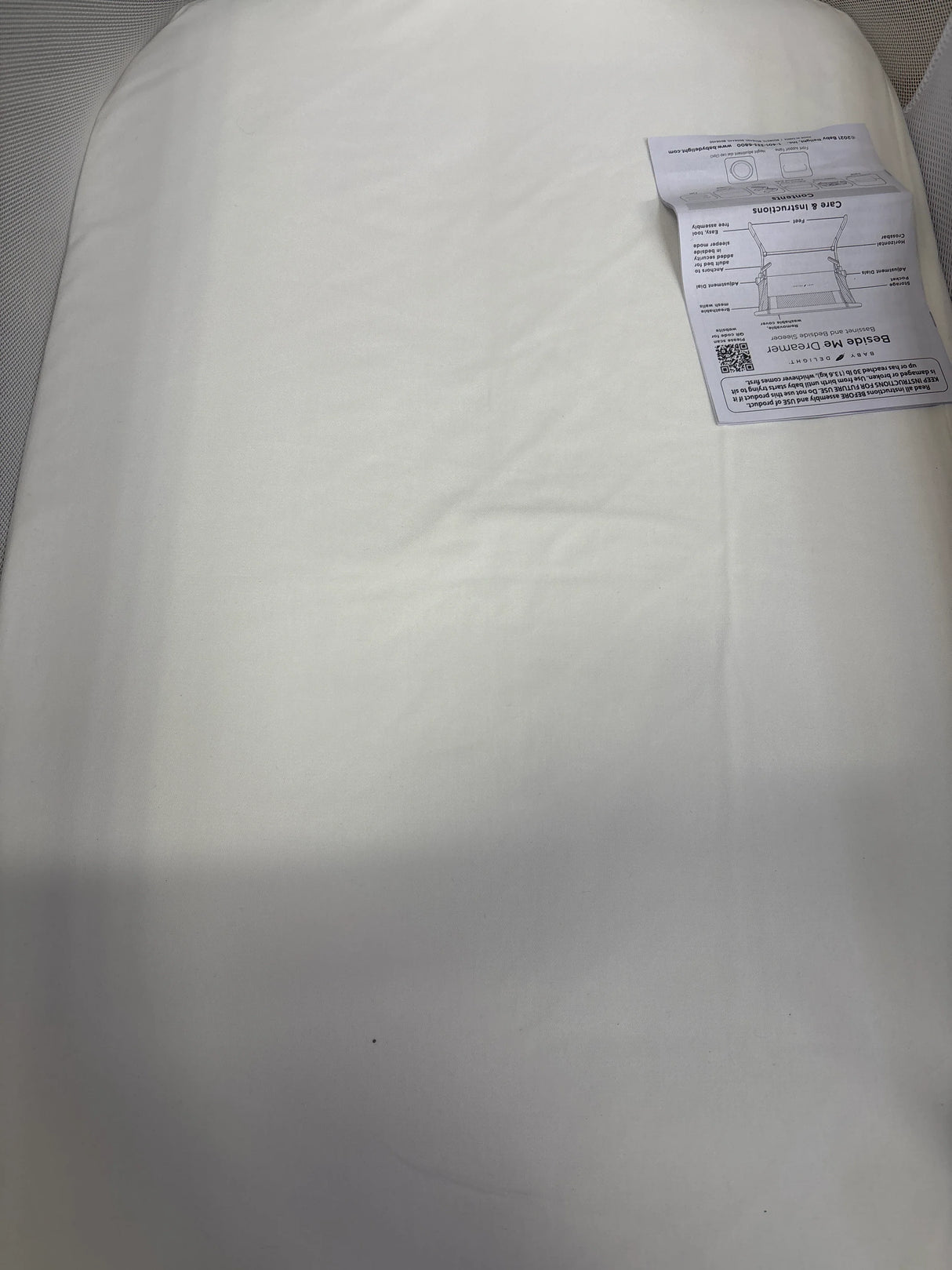 Top-down view of Baby Delight Beside Me Dreamer bedside bassinet showcasing mattress with white fitted sheet and instruction manual in corner